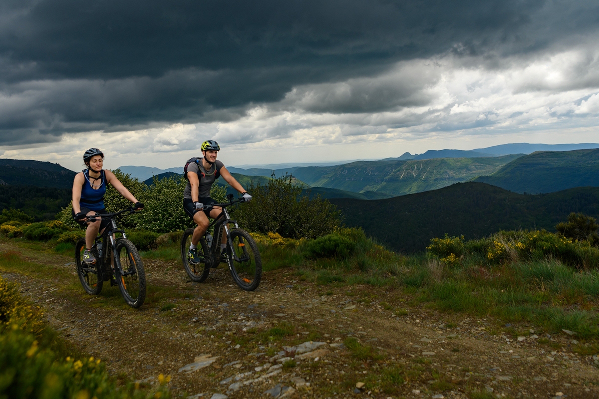 deux vététistes roulent sur un sentier avec des fleurs jaunes et les Cévennes en arrière plan, le ciel est menaçant, l'orage arrive